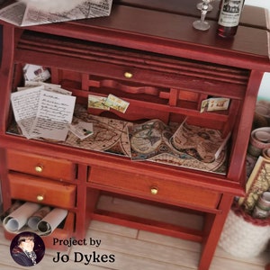 May include: A miniature wooden desk with a roll-top lid, drawers, and a map on the surface. The desk is decorated with vintage postcards and letters. The text "Project by Jo Dykes" is visible in the bottom right corner.