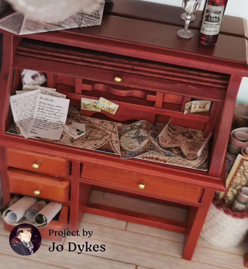 May include: A miniature wooden desk with a roll-top lid, drawers, and a map on the surface. The desk is decorated with vintage postcards and letters. The text "Project by Jo Dykes" is visible in the bottom right corner.