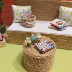 Op de afbeelding: Miniatuur buitensc&egrave;ne met een geweven bank, kussens en een ronde tafel. De tafel toont drie kleine potjes met de labels "Granny", "Mint Tea" en "Coconut", plus een tijdschrift. Een mand met tijdschriften en boeken staat op de bank.