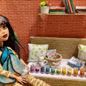 Op de afbeelding: Een pop in een turquoise en gouden outfit zit in de buurt van een miniatuur bank met kussens en een mand. Kleine potjes met verschillende geuren staan ervoor op een rij. Een miniatuur theepot en boeken zijn ook aanwezig. De achtergrond is een bakstenen muur.