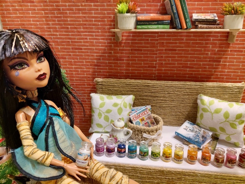 Op de afbeelding: Een pop in een turquoise en gouden outfit zit in de buurt van een miniatuur bank met kussens en een mand. Kleine potjes met verschillende geuren staan ervoor op een rij. Een miniatuur theepot en boeken zijn ook aanwezig. De achtergrond is een bakstenen muur.