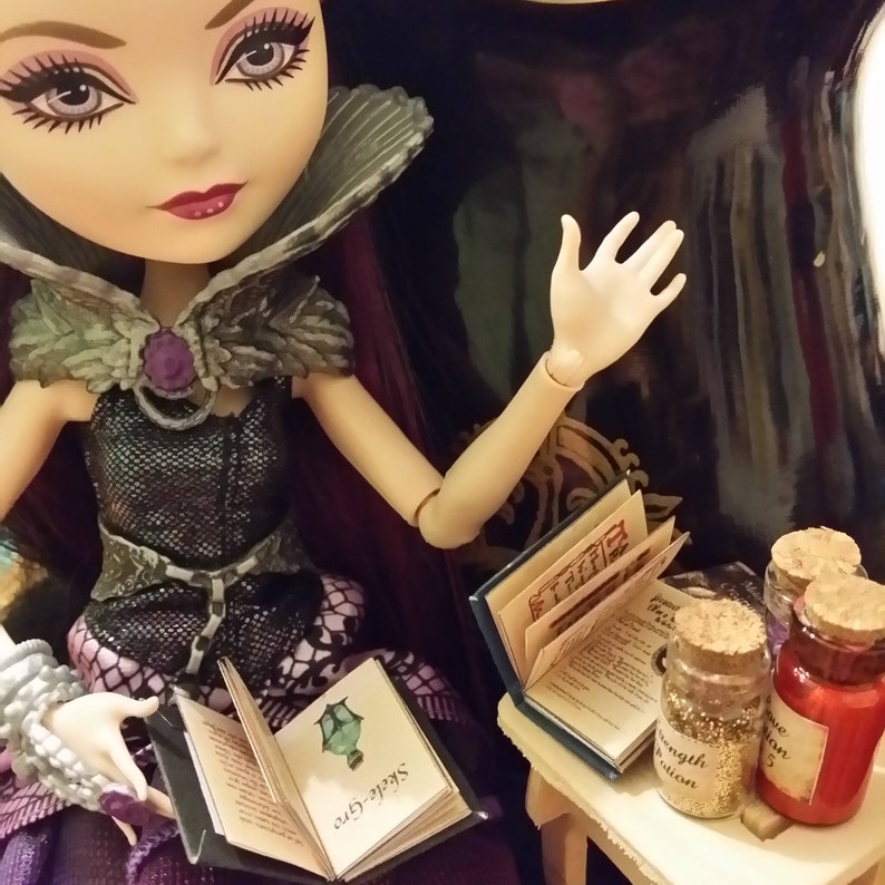 以下が含まれることがあります： 紫色の髪と銀と黒のドレスを着た人形が、「Seek the One」と書かれた表紙の開いた本を持っています。人形は椅子に座っており、その前に小さなテーブルがあります。テーブルの上には本と小さな瓶が3つ置かれています。瓶には、「Strength Potion」、「Love Potion」、「Invisibility Potion」とラベルが貼られています。
