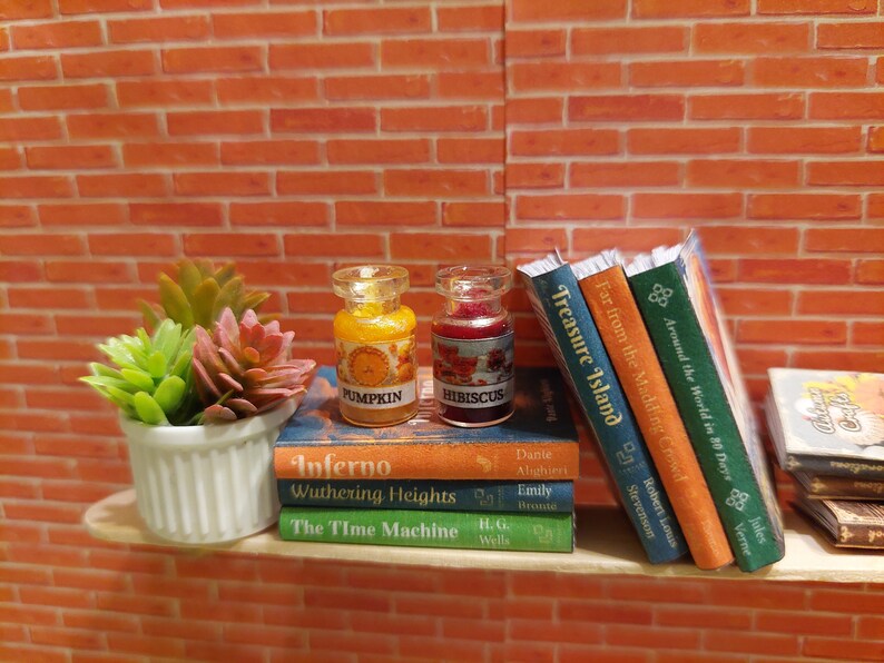Op de afbeelding: Miniatuur plank met boeken en decoratieve items. Twee kleine glazen potjes met de tekst "Pumpkin" en "Hibiscus" staan op een stapel boeken. Een kleine witte pot met vetplanten staat links. De achtergrond is een bakstenen muur.