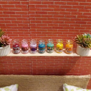 Op de afbeelding: Een miniatuurdisplay met zeven kleine glazen potjes met geuren als hibiscus, rozen, lavendel en pompoen. Elk potje is gelabeld en gevuld met kleurrijke inhoud. De potjes staan op een houten plank met pot-vetplanten tegen een bakstenen muur.