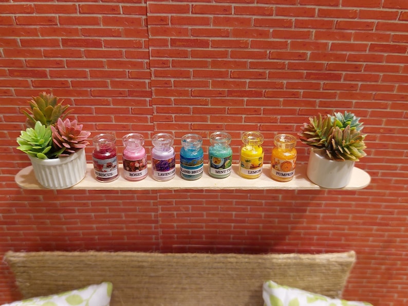 Op de afbeelding: Een miniatuurdisplay met zeven kleine glazen potjes met geuren als hibiscus, rozen, lavendel en pompoen. Elk potje is gelabeld en gevuld met kleurrijke inhoud. De potjes staan op een houten plank met pot-vetplanten tegen een bakstenen muur.