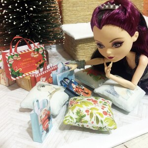 May include: A doll with purple hair and a black dress is surrounded by miniature Christmas shopping bags and pillows. The bags feature festive designs and the text "Merry Christmas". A small Christmas tree is in the background.