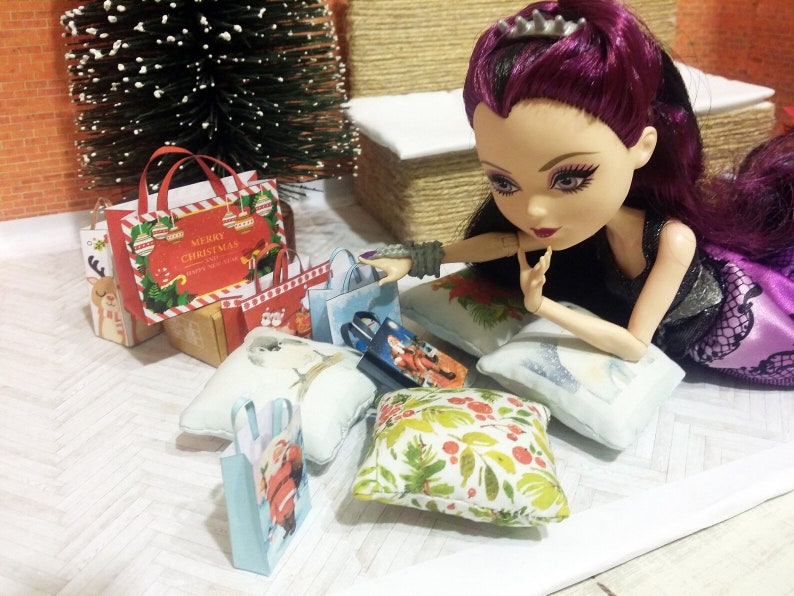 May include: A doll with purple hair and a black dress is surrounded by miniature Christmas shopping bags and pillows. The bags feature festive designs and the text "Merry Christmas". A small Christmas tree is in the background.