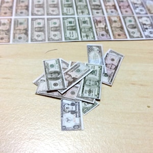 May include: A collection of miniature paper currency, including $100 and $5 bills, scattered on a light wooden surface. Some bills are stacked, while others are arranged in rows in the background. The bills feature portraits and green and gray details.