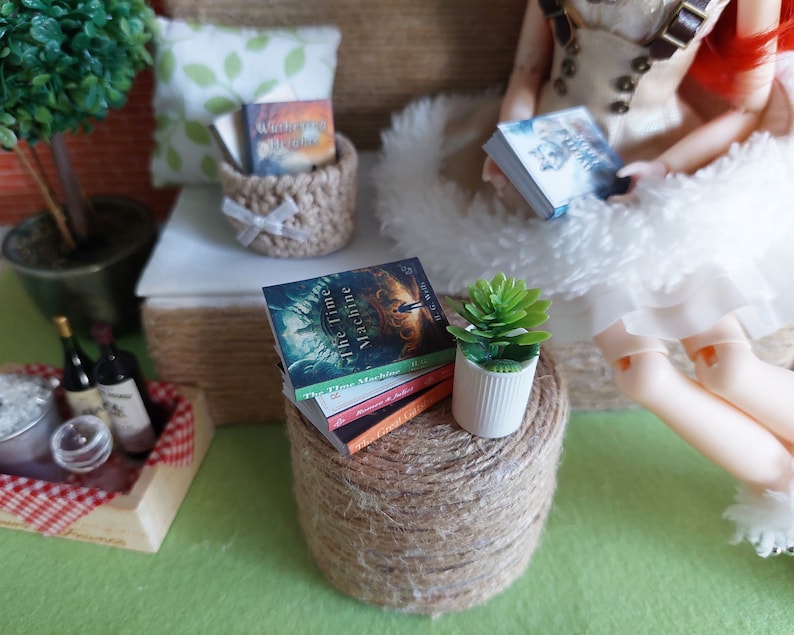 Op de afbeelding: Een miniatuur poppenhuis sc&egrave;ne met een kleine tafel, een stapel boeken, een plant in een pot en een pop die een witte jurk draagt. De boeken zijn getiteld "The Time Machine" en "The Time Machine II".