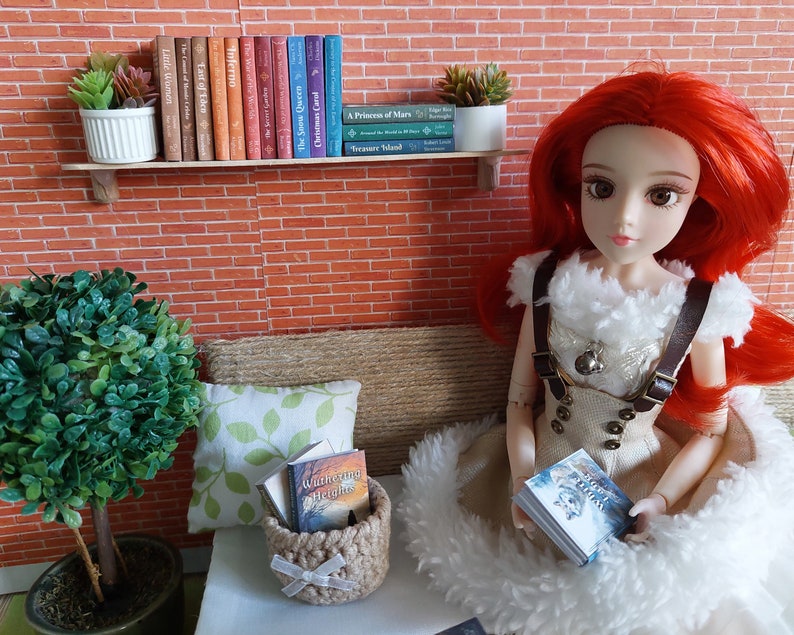 Op de afbeelding: Een roodharige pop met een witte en bruine jurk zit op een bank met een boek in haar handen. De pop is omringd door boeken en planten. De boeken staan op een plank boven de bank. De boeken zijn getiteld "Little Women", "East of Eden", "Inferno", "The Snow Queen", "Christmas Carol", "The Prince of Mars", "Treasure Island", en "Wuthering Heights".