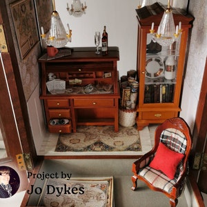 May include: A miniature dollhouse room with a wooden desk, a chair, a bookcase, and a map on the floor. The room is decorated with a chandelier and other miniature furniture. The text "Project by Jo Dykes" is visible in the bottom left corner of the image.