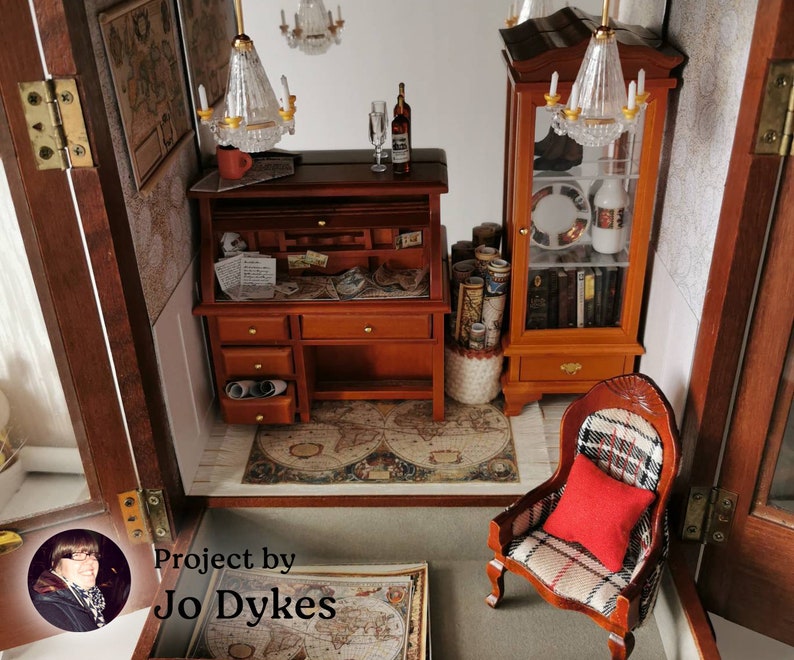 May include: A miniature dollhouse room with a wooden desk, a chair, a bookcase, and a map on the floor. The room is decorated with a chandelier and other miniature furniture. The text "Project by Jo Dykes" is visible in the bottom left corner of the image.