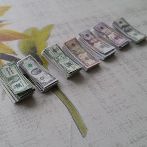 May include: Stacks of miniature United States dollar bills in various denominations, including one, five, ten, twenty, fifty, and one hundred dollar bills. The bills are arranged on a patterned surface with a floral design.