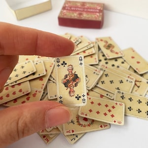 May include: A deck of playing cards with a Queen of Clubs card in the foreground. The Queen is depicted in a traditional style, wearing a crown and holding a scepter. The cards are in a vintage style with a cream background and black and red markings.