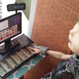 May include: A miniature black cash register with a screen displaying a fruit and vegetable selection. The screen shows a total of $43.50. The cash drawer is open, revealing neatly arranged paper money. A doll is reaching towards the cash register.