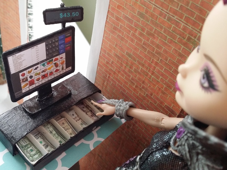 May include: A miniature black cash register with a screen displaying a fruit and vegetable selection. The screen shows a total of $43.50. The cash drawer is open, revealing neatly arranged paper money. A doll is reaching towards the cash register.