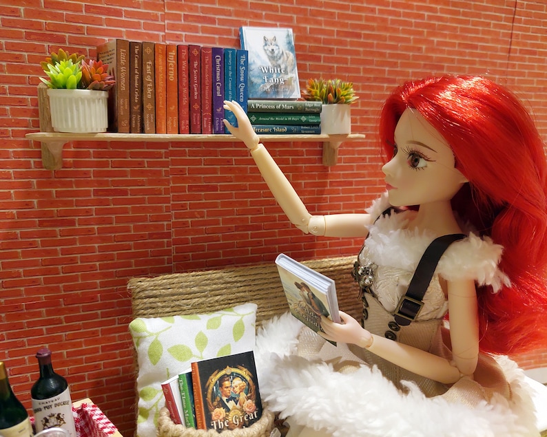 Op de afbeelding: Een pop met lang rood haar reikt naar een boek in een plank. De plank is gevuld met boeken met kleurrijke ruggen. De pop draagt een witte jurk met een bruine riem. De pop zit op een wit, pluche kussen. Er zijn twee flessen wijn en een glas op een rood-wit geruit tafelkleed.