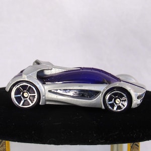 May include: A silver and blue toy car with a futuristic design. The car has a sleek, aerodynamic shape with a dark blue tinted windshield and silver wheels. The car is approximately 4 inches long.