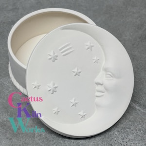 May include: White ceramic trinket box with a moon and stars design. The lid features a crescent moon with a smiling face and stars scattered around it. The box is perfect for storing small trinkets or jewelry.