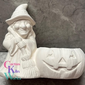 May include: A ceramic Halloween decoration featuring a smiling witch holding a broom next to a carved pumpkin. The witch wears a pointed hat and robe. The text "Cactus Kiln Works" is visible on the base.