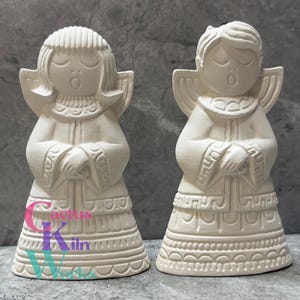 May include: Two off-white ceramic angel figurines with closed eyes and open mouths, as if singing. They have detailed wings, robes, and decorative bases. The figurines are unpainted and ready for customization. The logo "Cactus Kiln Works" is visible.