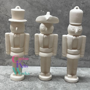 May include: Three white ceramic nutcracker figurines. The nutcracker on the left is wearing a tall hat. The nutcracker in the middle is wearing a hat with a brim. The nutcracker on the right is wearing a tall hat with a brim.
