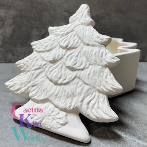 May include: A white ceramic Christmas tree-shaped container with a detailed, textured design. The container has a matching white, rectangular base with a wavy top edge. The logo "Cactus Kiln Works" is visible on the base.