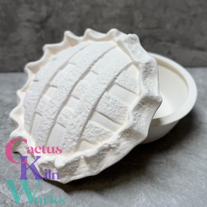 May include: A white ceramic pie dish with a textured, lattice-patterned lid. The lid is slightly ajar, revealing the dish's interior. The dish has a scalloped edge. The text "cactus Kiln Works" is visible in the lower left corner.