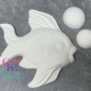 May include: A white ceramic fish with detailed fins and scales, accompanied by two white spherical objects. The fish has a smiling expression. The image includes the text "Creative Kiln Works" in the lower left corner.