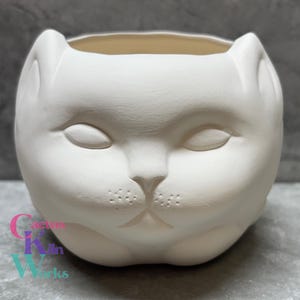 May include: White ceramic planter shaped like a cat's face. The planter has closed eyes, a small nose, and a slightly downturned mouth.