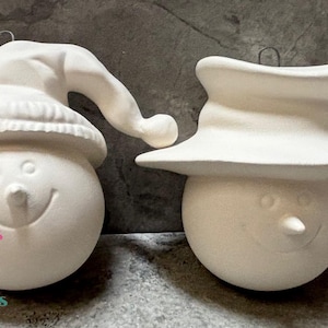 May include: Two unpainted ceramic snowman ornaments. One snowman wears a knit cap, the other a top hat. Both have smiling faces, button eyes, and carrot noses. Each ornament has a metal loop for hanging.