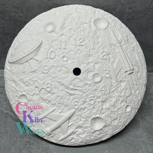 May include: A white ceramic clock face with a textured moon surface design. Features a star in the center, numbers, stars, a rocket, and a UFO. The clock face is round and has a hole in the center for the clock hands.