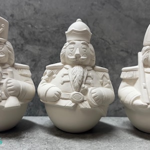 May include: Three unpainted ceramic nutcracker figurines. Each figure has a detailed uniform, hat, and a stern expression. The figures are in a row, ready for painting. The Cactus Kiln Works logo is visible.