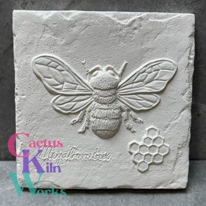 May include: A square, off-white stone tile with a detailed relief of a bee, wings spread, and a honeycomb pattern. The text "Megabombus" is below the bee. The tile is likely for decorative purposes.