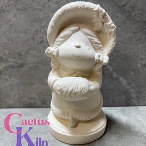 May include: A bisque ceramic figurine of a smiling girl wearing a bonnet and holding a fluffy object. The figurine is unpainted and ready for customization. The base of the figurine is round. The text "Cactus Kiln Works" is at the bottom.