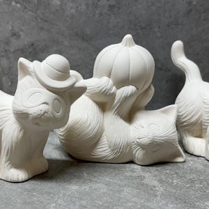 May include: Three unpainted ceramic cat figurines. One cat is holding a pumpkin, another is wearing a hat, and the third is wearing a witch hat. The figurines are white and ready to be painted.