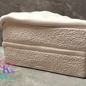 May include: A ceramic jewelry box shaped like a slice of cake. The off-white box has a textured surface and a curved top, resembling frosting. The box is rectangular with a horizontal band near the bottom. The logo "Canvas Kiln Works" is visible.