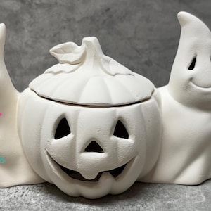 May include: White ceramic Halloween decoration featuring two ghosts flanking a pumpkin with a lid. The pumpkin has a carved jack-o'-lantern face.