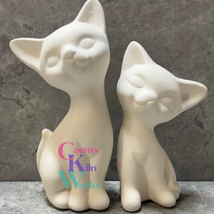 May include: Two unpainted ceramic cat figurines. The cats are in a sitting position with their eyes closed and a slight smile. The larger cat is taller than the smaller one. The text "Cactus Kiln Works" is printed on the larger cat.