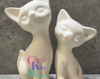 May include: Two unpainted ceramic cat figurines. The cats are in a sitting position with their eyes closed and a slight smile. The larger cat is taller than the smaller one. The text "Cactus Kiln Works" is printed on the larger cat.