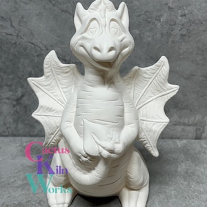 May include: A white ceramic dragon figurine with detailed wings, a smiling face, and a textured body. The dragon is in a seated position with its hands clasped. The text "Cactus Kiln Works" is visible on the base.