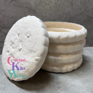 May include: A white ceramic container with a lid, resembling a stack of cookies. The lid is oval-shaped with small indentations, and the container has a ridged edge. The text "Cactus Kiln Works" is visible on the lid.