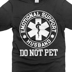 May include: Black t-shirt with a white graphic. The graphic features a white star of life symbol with a snake and staff, surrounded by the words "EMOTIONAL SUPPORT HUSBAND" and "DO NOT PET".