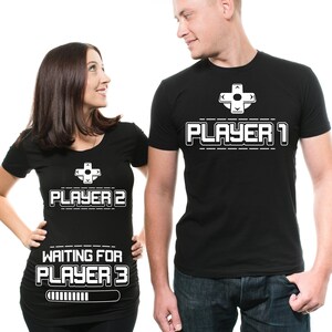 May include: A couple wearing matching black t-shirts with a video game theme. The woman's shirt says "Player 2 Waiting for Player 3" with a progress bar. The man's shirt says "Player 1".