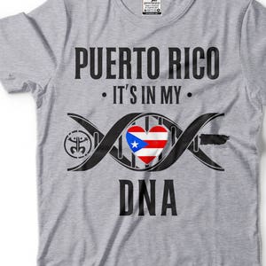 Puede incluir: Camiseta gris con texto negro que dice "Puerto Rico. Está en mi ADN." Una cadena de ADN negra con un corazón que contiene la bandera puertorriqueña está en el centro de la camiseta.