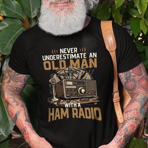 Könnte beinhalten: Ein schwarzes Kurzarm-T-Shirt mit einer Grafik eines alten Amateurfunkgeräts inklusive Antenne und Mikrofon. Der Text lautet: NEVER UNDERESTIMATE AN OLD MAN WITH A HAM RADIO. Das Design besteht aus beigen und weißen Buchstaben auf dem schwarzen Stoff.