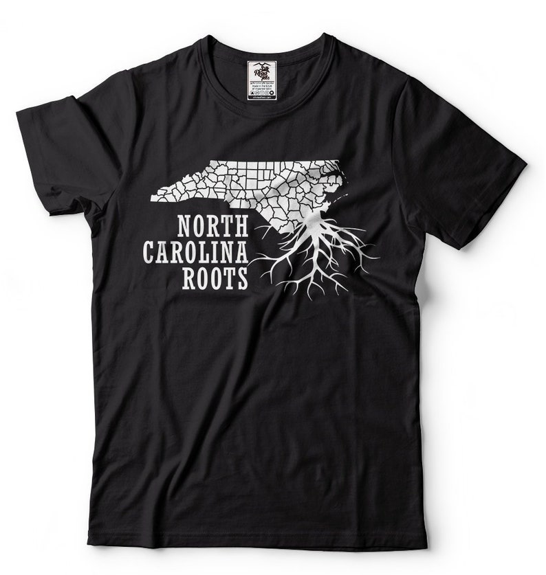 Puede incluir: Camiseta negra con un contorno blanco del estado de Carolina del Norte y ra&iacute;ces de &aacute;rbol. El texto "NORTH CAROLINA ROOTS" est&aacute; impreso debajo del contorno del estado.