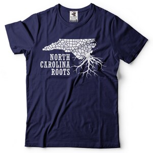 Puede incluir: Una camiseta azul marino con un contorno blanco del estado de Carolina del Norte y ra&iacute;ces de &aacute;rbol. El texto "NORTH CAROLINA ROOTS" est&aacute; impreso debajo del contorno del estado.