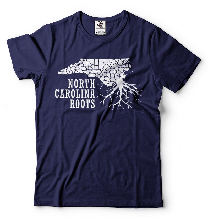Puede incluir: Una camiseta azul marino con un contorno blanco del estado de Carolina del Norte y ra&iacute;ces de &aacute;rbol. El texto "NORTH CAROLINA ROOTS" est&aacute; impreso debajo del contorno del estado.