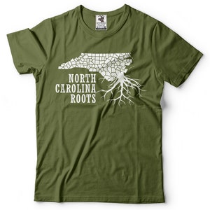 Puede incluir: Una camiseta verde con un contorno blanco del estado de Carolina del Norte y un dise&ntilde;o de ra&iacute;z de &aacute;rbol. El texto "North Carolina Roots" est&aacute; impreso debajo del contorno del estado.